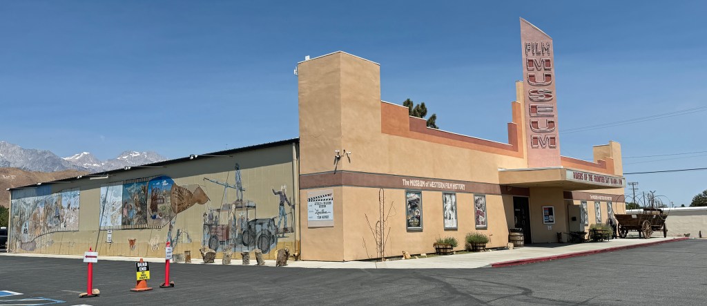 Exterior view of the Film Museum with murals depicting film history, surrounded by a parking lot and mountains in the background.