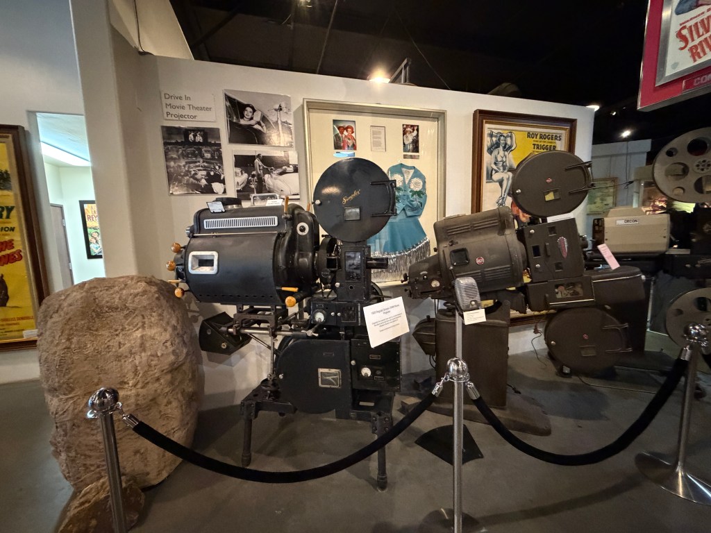 A vintage drive-in movie theater projector displayed in a museum, surrounded by ropes for crowd control, with historical photos and memorabilia on the walls.