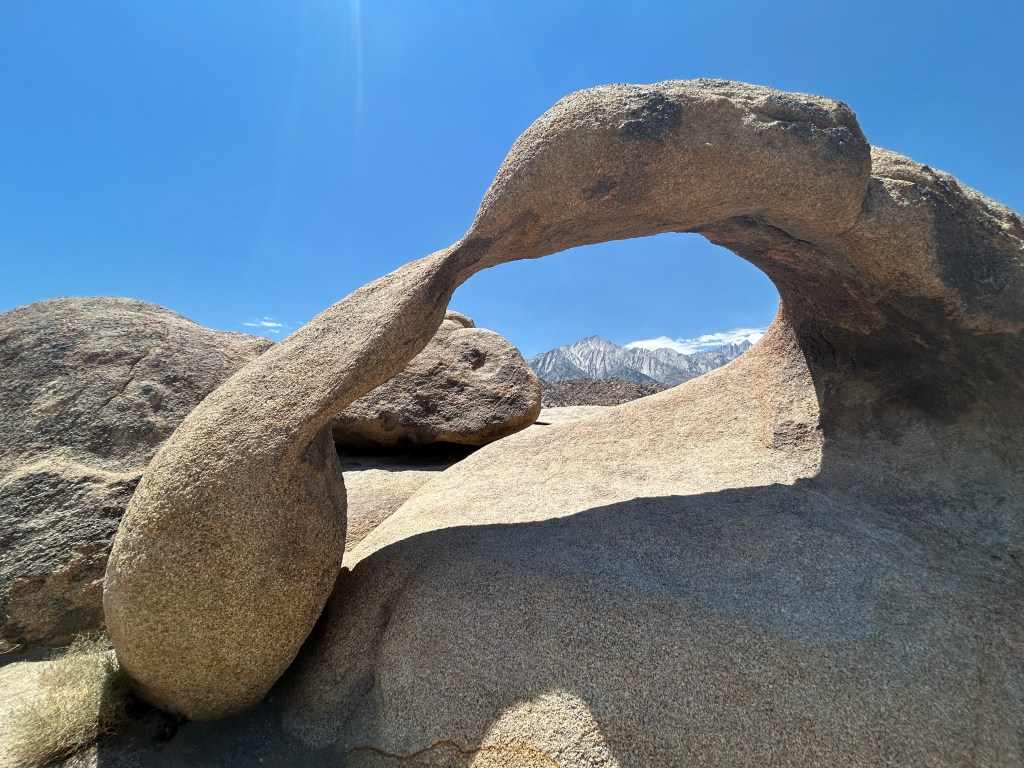 A unique rock formation creating a natural arch against a clear blue sky, with distant mountains visible in the background.