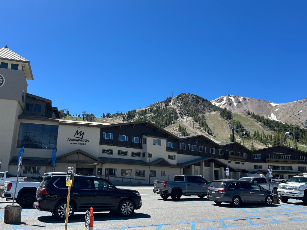 View of Mammoth Main Lodge with parked vehicles in the foreground and mountains in the background under a clear blue sky.